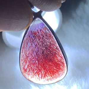 Puede incluir: Un colgante en forma de lágrima con una piedra transparente y facetada. La piedra tiene un color rojo y morado con un patrón estriado. El colgante está hecho de plata.