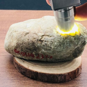 May include: A close-up of a light green stone being illuminated by a UV light. The stone is resting on a wooden base. The stone has a red inscription that reads "00514 1372".