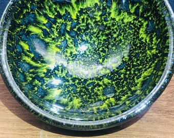 May include: A small, round ceramic bowl with a black and green speckled glaze. The bowl has a glossy finish and a slightly raised rim.