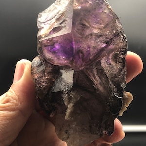May include: A raw amethyst crystal cluster with a deep purple center and translucent, layered edges. The natural gemstone displays a mix of dark and light tones, with a rough, textured surface. The crystal is held in a hand against a dark background.