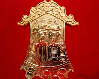 May include: A gold-colored, bell-shaped pendant with intricate embossed designs and Chinese characters. The pendant has a loop at the top for hanging and is set against a red background. The design includes floral and geometric patterns.