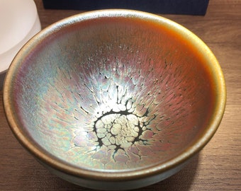 May include: A small, round, ceramic teacup with a brown, iridescent glaze. The glaze has a crackled effect, creating a unique pattern of light and dark areas.