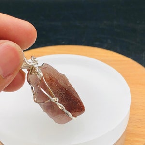 May include: A reddish-brown crystal pendant wrapped in silver wire. The pendant is displayed on a white circular surface.