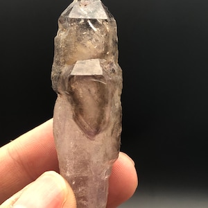 May include: A raw, natural amethyst crystal with a pointed tip and a rough, textured surface. The crystal is a translucent purple color with darker inclusions and is held against a dark background.