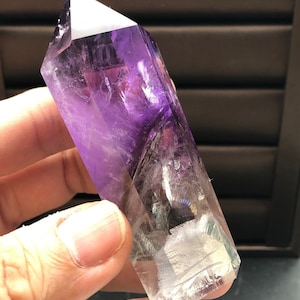 Puede incluir: Una punta de cristal de amatista transparente con bandas púrpuras y blancas. El cristal es puntiagudo en la parte superior y tiene una base plana.