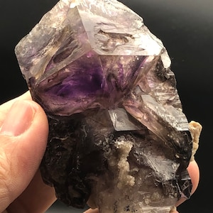 May include: A raw amethyst crystal cluster with a deep purple center and translucent, light purple edges. The natural gemstone has a rough, textured surface and is held in a hand against a dark background.
