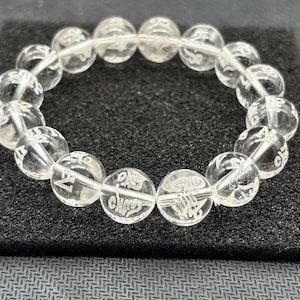 May include: Clear quartz crystal bracelet with etched symbols. The bracelet features round, translucent beads strung together, each displaying intricate, etched designs. The bracelet is displayed on a black surface.