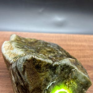 Puede incluir: Una piedra en bruto de color verde oliva con una superficie natural y texturizada. Una luz verde brillante brilla a través de una parte de la piedra, resaltando su cualidad translúcida. La piedra está sobre un fondo oscuro.