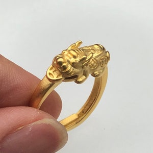 May include: A gold ring featuring a dragon design. The dragon is facing forward with its wings outstretched. The ring is crafted from smooth, shiny metal.
