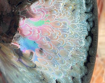 Könnte beinhalten: Nahaufnahme einer Keramikschale mit einer einzigartigen, irisierenden Glasur. Das Innere der Schale weist eine Mischung aus Farben auf, darunter Rosa, Blau und Grün, mit einem strukturierten, organischen Muster. Der Rand ist dunkel und kontrastiert mit der lebendigen Mitte.