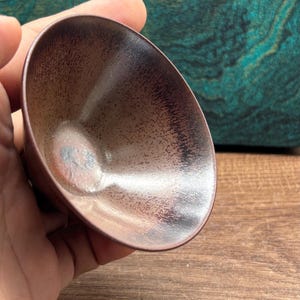May include: A small, brown ceramic bowl with a speckled interior and a smooth, glossy exterior. The bowl is held in a hand, with a wooden surface in the background. The bowl's shape is oval and it has a dark rim.