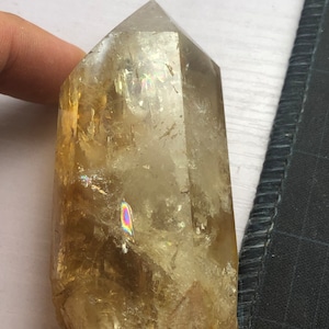 Pode incluir: Uma ponta de cristal de citrino dourado transparente e facetado com inclusões arco-íris iridescentes. O cristal tem aproximadamente 10 cm de altura e tem uma forma hexagonal.