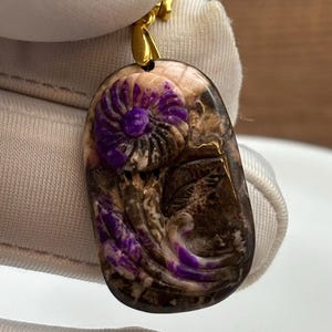 Puede incluir: Un colgante de piedra p&uacute;rpura y marr&oacute;n con un dise&ntilde;o de flor tallada. El colgante est&aacute; unido a una cadena de oro.