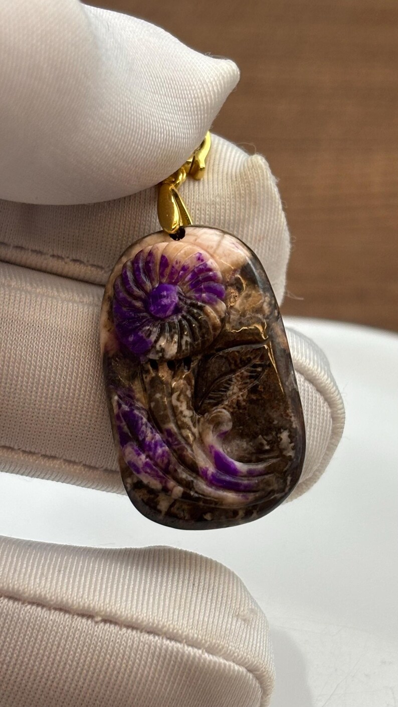 Puede incluir: Un colgante de piedra p&uacute;rpura y marr&oacute;n con un dise&ntilde;o de flor tallada. El colgante est&aacute; unido a una cadena de oro.