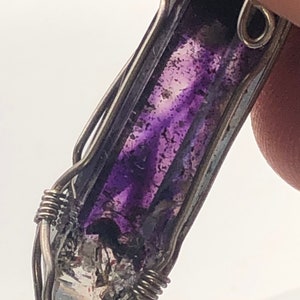 May include: A purple and clear crystal pendant wrapped in silver wire. The crystal has a pointed end and is set in a wire cage.