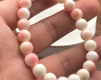 May include: A bracelet made of round, polished beads in shades of pink and white. The beads are strung together to form a complete circle. The bracelet is held in a hand, showcasing the color and texture of the beads.