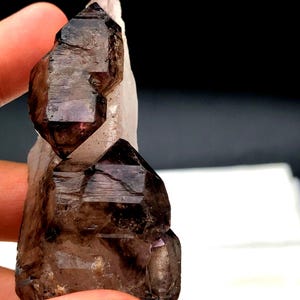 May include: A smoky quartz crystal cluster with a dark brown to purple hue. The crystal has a pointed shape and is translucent.