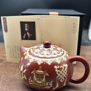 Puede incluir: Una tetera de cerámica roja con diseños florales y geométricos blancos y dorados. La tetera tiene una tapa con un pomo en la parte superior. La tetera está sobre una superficie de madera.