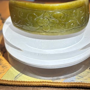 Puede incluir: Un brazalete de jade verde oliva curvado con detalles florales y arremolinados tallados. El brazalete se exhibe sobre un soporte circular blanco. El diseño del brazalete presenta un motivo floral central y patrones fluidos.