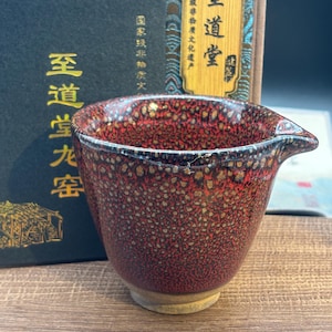 May include: A red and brown speckled ceramic tea cup with a spout. The cup has a wide opening and a slightly flared base. The background includes a box with Chinese characters.