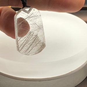 May include: A clear quartz pendant with silver rutilated inclusions. The pendant is shaped like a teardrop and has a black cord.