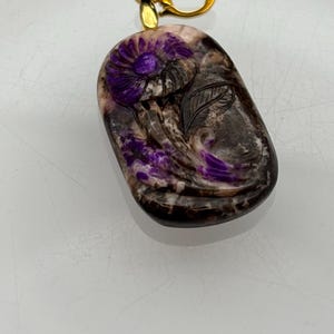 Puede incluir: Un colgante de piedra tallada de color morado y blanco con un dise&ntilde;o floral y una anilla de oro.