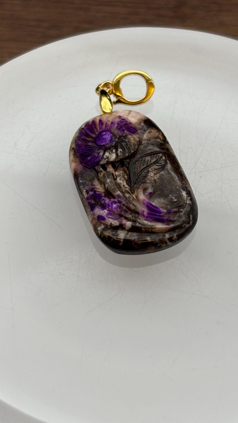 Puede incluir: Un colgante de piedra tallada de color morado y blanco con un dise&ntilde;o floral y una anilla de oro.