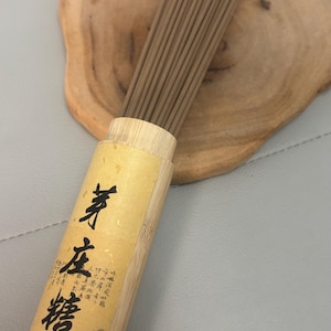 Peut inclure: Un ensemble de bâtonnets d'encens bruns dans un contenant en bambou avec des caractères chinois. Le contenant cylindrique a une étiquette jaune avec du texte noir et une peinture de paysage. Posé sur une tranche de bois.