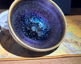 Puede incluir: Un pequeño cuenco de té de cerámica con un interior profundo e iridiscente. El interior del cuenco presenta un patrón arremolinado de azules, morados y toques dorados. El borde es de un marrón cálido y terroso. Una caja azul oscuro con borla está en el fondo.