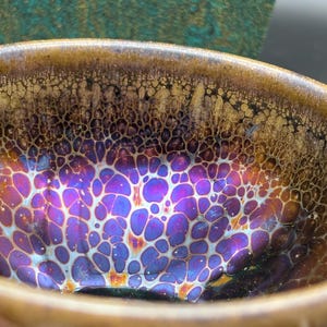 Pode incluir: Close-up de uma tigela de cerâmica com um esmalte iridescente único. O interior apresenta uma exibição vibrante de roxos, azuis e dourados, criando um padrão celular. A borda é marrom quente, adicionando contraste. Uma peça decorativa.