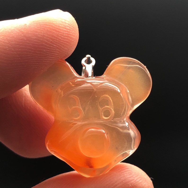 K&ouml;nnte beinhalten: Ein durchscheinender, orangefarbener Anh&auml;nger in Form einer Zeichentrickmaus. Der Anh&auml;nger hat oben eine silberfarbene Schlaufe zum Aufh&auml;ngen. Das Gesicht der Maus ist mit Augen, Nase und Ohren detailliert. Der Anh&auml;nger wird zwischen zwei Fingern vor dunklem Hintergrund gehalten.