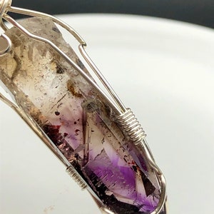 May include: A silver wire wrapped pendant featuring a purple and white amethyst crystal with inclusions. The crystal is faceted and has a pointed tip.