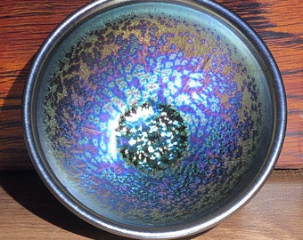 Könnte beinhalten: Eine kleine, irisierende Teeschale mit einem metallisch silbernen Rand. Das Innere weist ein wirbelndes Muster aus Blau-, Grün- und Goldtönen auf. Die Schale steht vor einer Holzkiste mit grauem Stofffutter, was auf eine Geschenkbox hindeutet.
