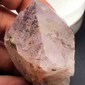 May include: A clear quartz crystal with a pointed tip. The crystal has a light purple hue with dark speckling and some green inclusions. The crystal is held in a hand.