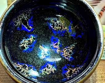 May include: A ceramic bowl with a dark blue interior, featuring a speckled, iridescent pattern of blue, yellow, and gold. The rim has a gold metallic finish. The bowl is likely handmade and suitable for serving food.