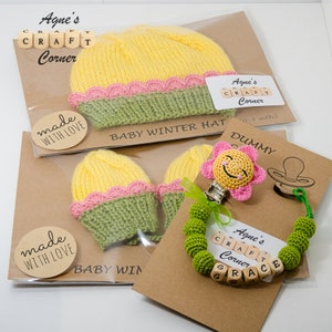 Könnte beinhalten: Ein handgemachtes Baby-Set in Gelb, Grün und Rosa. Das Set beinhaltet eine gestrickte Mütze, Fäustlinge und einen gehäkelten Schnullerclip mit einem Blumenanhänger. Der Schnullerclip hat Holzperlen, die den Namen "Grace" buchstabieren.