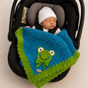 Könnte beinhalten: Ein schlafendes Neugeborenes in einem Autositz, eingehüllt in eine handgefertigte blau-grüne Häkeldecke. Die Decke hat ein Frosch-Applikation mit einer grünen Schleife. Das Baby trägt eine weiße Mütze.