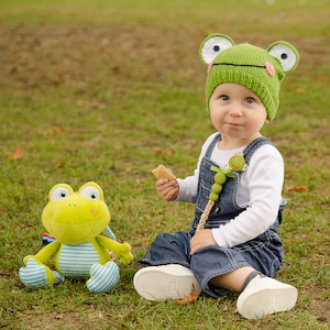 Könnte beinhalten: Ein Kind trägt einen grünen Froschhut mit großen weißen Augen und rosa Akzenten, kombiniert mit einer Jeans-Latzhose und einem weißen Langarmshirt. Ein Plüschfroschspielzeug mit blau-weiß gestreiften Akzenten liegt daneben. Ein Schnullerclip mit dem Namen "DOMINIK" ist befestigt.