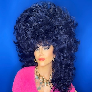 Könnte beinhalten: Eine Person mit einer großen, dunkelblauen, lockigen Perücke, die einen rosa, flauschigen Cardigan mit tiefem Ausschnitt und einer goldenen Kettenkette trägt. Die Person hat Tätowierungen auf ihren Armen und ihrer Brust.