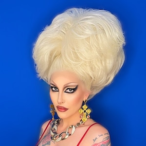 Könnte beinhalten: Eine Person mit einer großen blonden Bouffant-Frisur trägt ein rotes Tanktop und goldenen Schmuck. Die Person hat Tätowierungen auf ihren Armen, darunter einen roten Teufel, ein Auto und die Worte "love, I'm not sure I can leave, I'm not sure I want to".