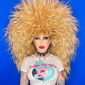 Könnte beinhalten: Eine Frau mit einer großen, blonden, flauschigen Perücke trägt ein weißes Crop-Top mit dem Text "Truffle Pig Wig" und eine schwarze Halskette mit einem silbernen Ring. Sie trägt eine schwarze Hose und hat Tattoos auf ihren Armen.