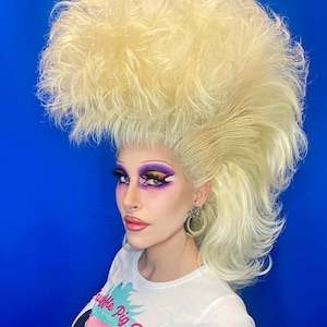 May include: A person with a large blonde wig styled in a high, teased updo. They are wearing a white t-shirt with the text "Ruffle Pig Wigs" and a graphic of a pig with a large, ruffled wig.