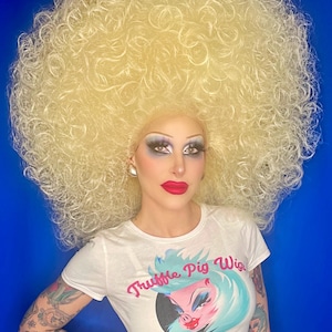 Może przedstawiać: Osoba w białym crop topie z napisem "Truffle Pig Wigs" i dużą blond, kręconą peruką. Osoba ma tatuaże na ramionach.