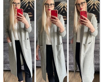 May include: A light gray long cardigan with two front pockets. The cardigan is worn over a white top and black pants. The cardigan is knee-length and has a relaxed fit. The outfit is completed with white sneakers.