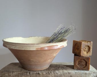 May include: A terracotta mixing bowl with a cream-colored interior glaze containing a bundle of dried lavender sprigs, positioned next to a stack of two square, amber-colored soap bars featuring a hexagonal embossed design on their front faces.