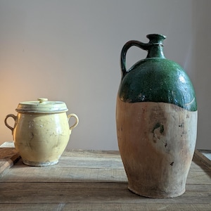 May include: Two antique ceramic vessels on a wooden surface. A small, yellow lidded pot with handles sits next to a large, green-topped jug with a handle. The jug has a light brown base. The items appear aged and rustic.