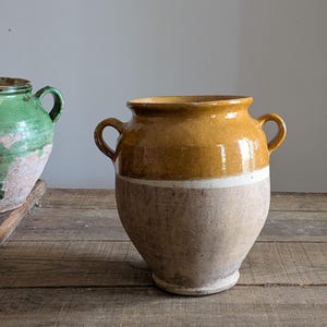 以下が含まれることがあります： 黄色い上部とタン色の底部を持つ、2つのハンドルが付いた大きなアンティークセラミックポット。2色を分ける白い帯があります。左側には別の緑色のポットが部分的に見えます。ポットは木製の表面にあります。