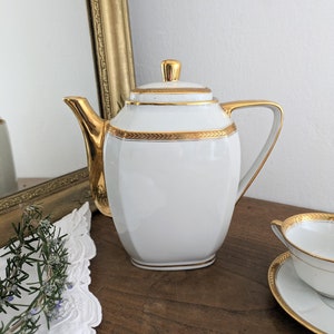 Op de afbeelding: Witte porseleinen theepot met gouden randen en details. De theepot heeft een geometrische vorm, een deksel en een handvat. Een bijpassende kop en schotel zijn zichtbaar. Onderdeel van een theeservies.