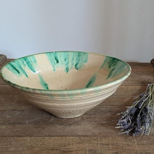 May include: A large, cream-colored ceramic bowl with a green rim and streaks. The bowl sits on a wooden surface next to a bundle of dried lavender. The bowl has a wide, shallow shape.