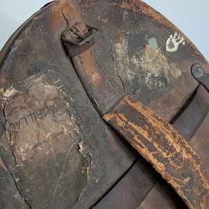 Pu&ograve; includere: Una cappelliera in pelle marrone usurata con manico logoro, fibbia in metallo e due etichette di carta strappate. Un'etichetta mostra chiaramente il testo AURILLAC e Gare destinataire, l'altra mostra testo parziale incluso GES PL.
